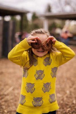 A little girl dressed in a fall sweater covers in owls holds 2 apple cider donuts over her eyes at Great Country Farms apple picking festival